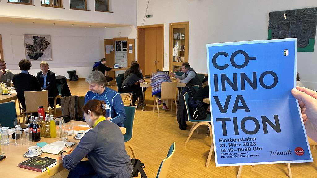 Menschen an Tischen arbeitend, im Vordergrund ein Plakat mit der Aufschrift Co-Innovation