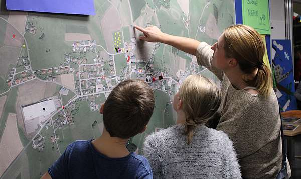 Zwei Kinder und eine Frau stehen vor einer Landkarte, die auf einer Pinnwand klebt.