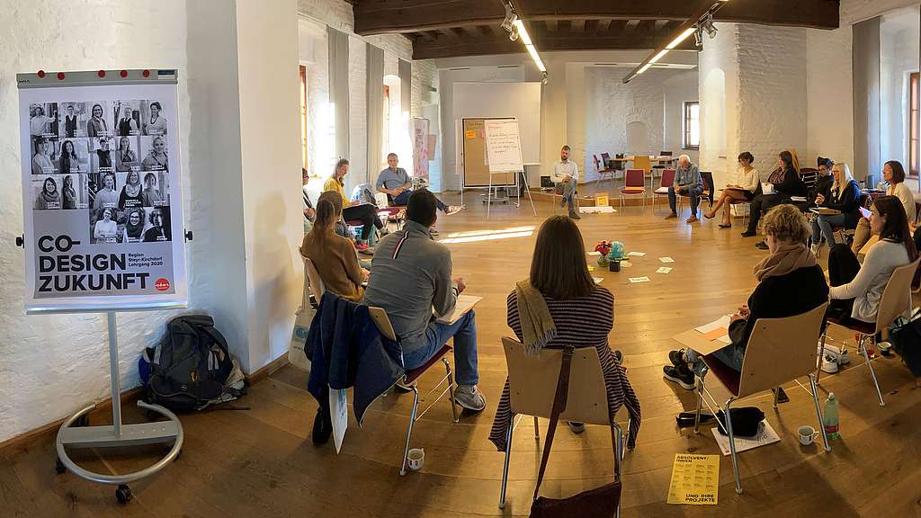 20 TeilnehmerInnen auf Sesseln in einem Kreis sitzend, im Vordergrund ein Flipchart mit Aufschrift Co-Design Zukunft
