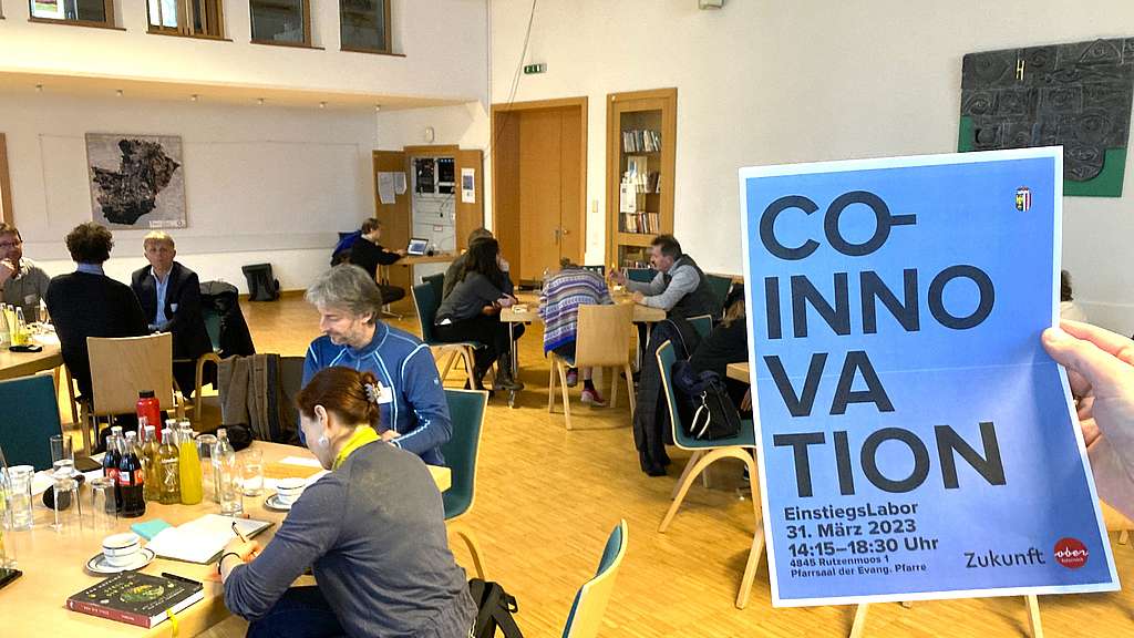 Menschen, sitzen arbeitend an Tischen, im Vordergrund ein Plakat mit der Aufschrift "Co-Innovation"