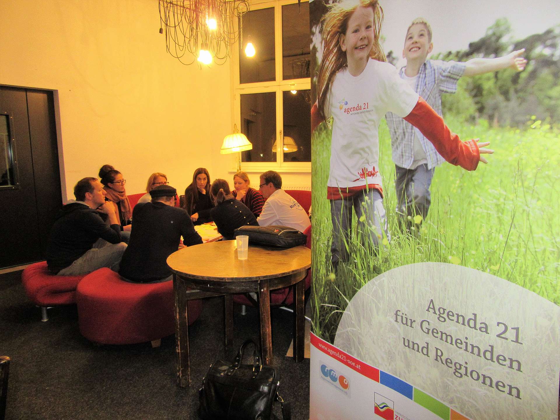 Sieben Personen sitzen an einem Tisch, im Vordergrund ein Roll-Up mit der Aufschrift Agenda 21 Oberösterreich